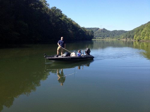 A person fishing from a small boat on a calm lake surrounded by green hills and trees.