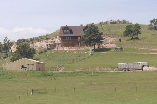 A scenic view of a house on a hill, with a barn and trailer in a green pasture under a clear sky.