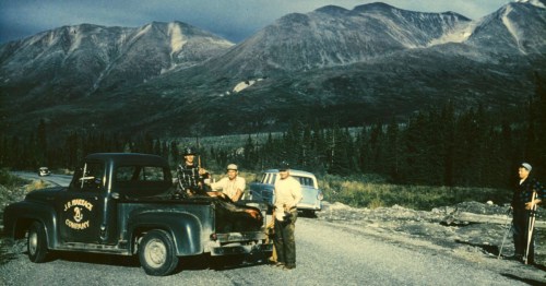 A vintage truck with four people stands on a gravel road, surrounded by mountains and trees under a cloudy sky.