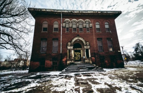 Abandoned red brick building with a grand entrance, snow on the ground, and graffiti reading "Know Thy Self."