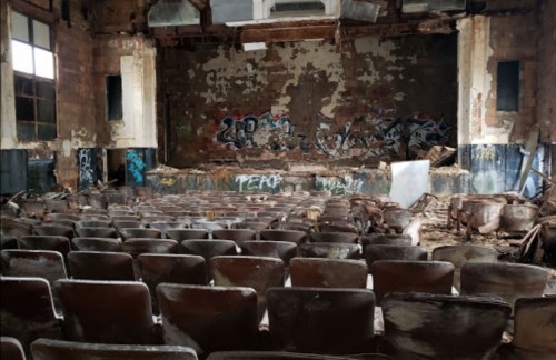 Abandoned theater interior with broken seats, graffiti on walls, and debris scattered across the stage.