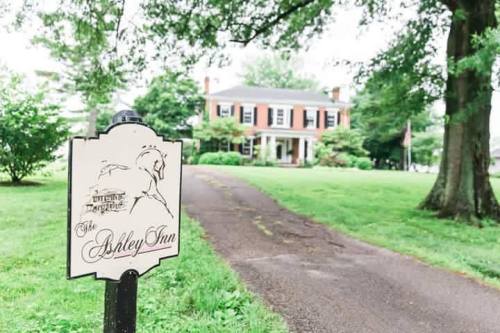 A sign for "The Ashley Inn" in front of a large, historic house surrounded by greenery.