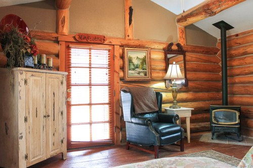 Cozy log cabin interior featuring a blue armchair, wooden cabinet, lamp, and a small wood stove by a door.