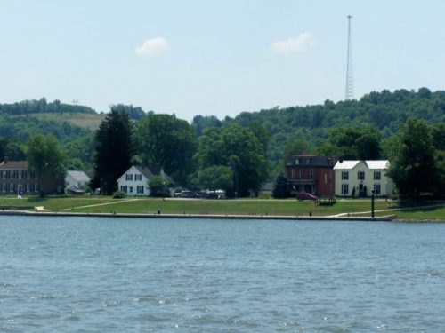 A serene lakeside view featuring houses, green trees, and a distant radio tower under a clear blue sky.