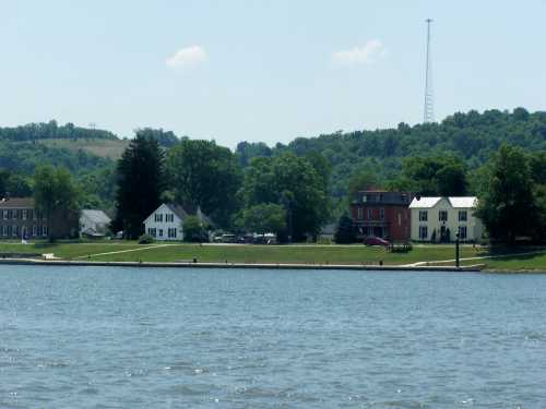 A serene lakeside view featuring houses, trees, and a distant hill with a radio tower under a clear blue sky.