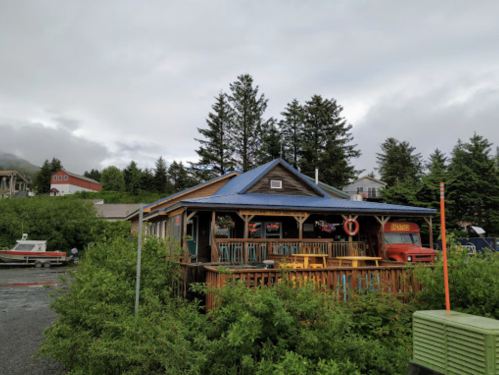 A rustic wooden building with a blue roof, surrounded by greenery and boats, under a cloudy sky.