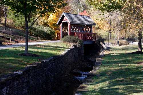 A small red covered bridge over a stream, surrounded by trees with autumn foliage in a peaceful park setting.