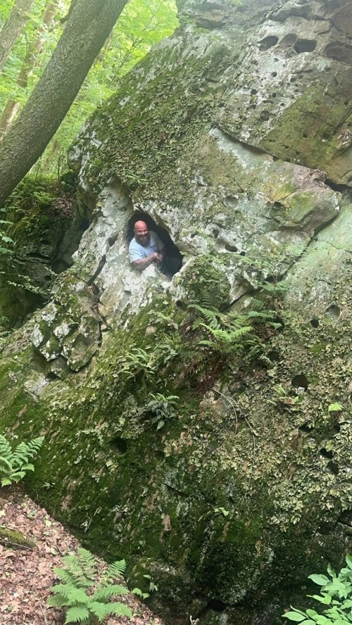 A person peeks out from a small opening in a moss-covered rock formation in a lush forest.