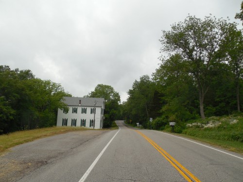 A winding road with a white house on the left, surrounded by greenery under a cloudy sky.