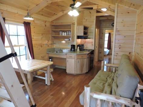 Cozy wooden cabin interior with a kitchen, dining table, and seating area, featuring warm lighting and rustic decor.