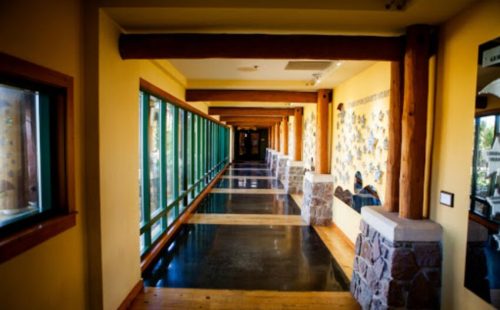 A long hallway with large windows, wooden beams, and stone accents, leading to a well-lit area.