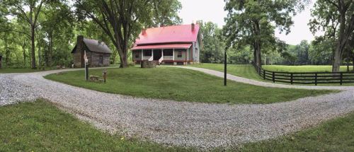 A scenic view of a gravel driveway leading to a house with a red roof, surrounded by trees and a rustic cabin nearby.