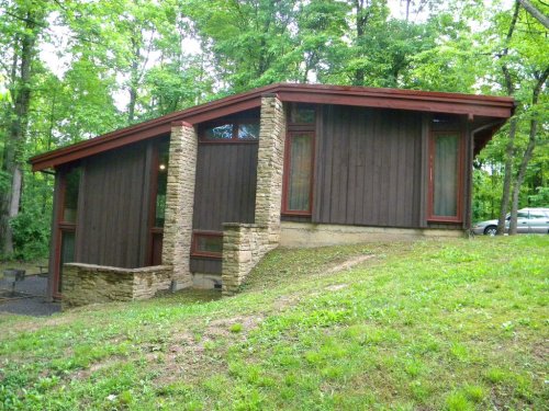 A modern wooden house with stone accents, set on a grassy slope surrounded by trees.