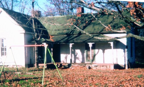A weathered house with a green roof, a swing set in the foreground, and fallen leaves scattered on the ground.