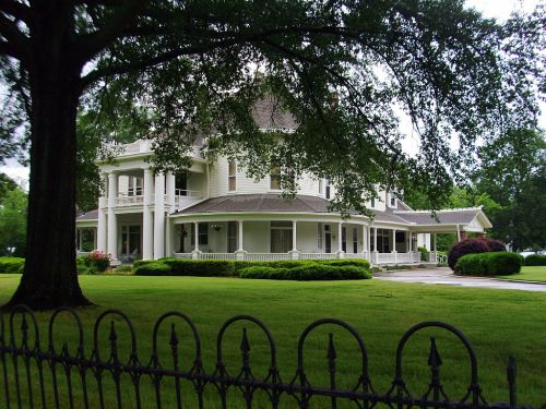 A large, elegant white house with a wraparound porch, surrounded by green grass and trees, viewed through a decorative fence.