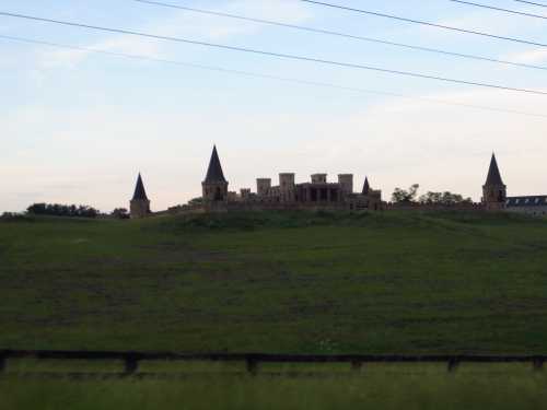 A large, unfinished castle with towers sits atop a green hill under a cloudy sky, surrounded by power lines.