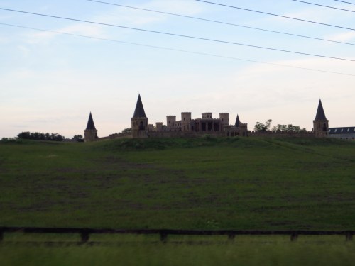 A large, unfinished castle with towers sits atop a green hill under a cloudy sky, surrounded by power lines.