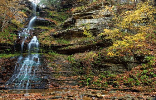 A serene waterfall cascading down rocky cliffs, surrounded by autumn foliage and a forested landscape.