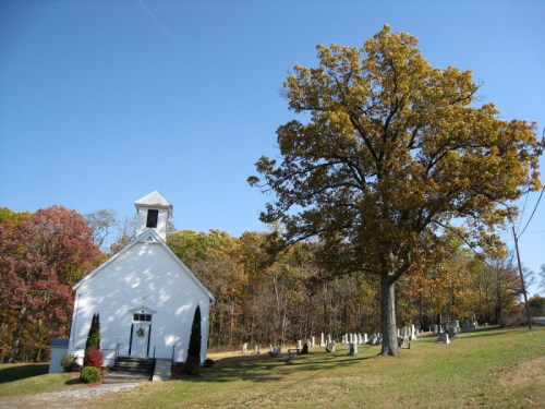 A white church beside a large tree and a cemetery, set against a clear blue sky and autumn foliage.