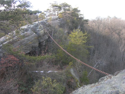 A long chain bridge stretches across a rocky cliff, surrounded by trees and a forested landscape.
