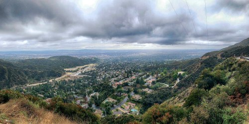 A panoramic view of a valley with houses, surrounded by hills under a cloudy sky.