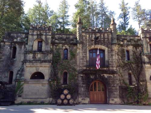 A stone castle-like building surrounded by trees, featuring turrets and an American flag, with barrels in front.
