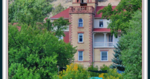 Historic building with a tower, surrounded by lush greenery and colorful flowers, set against a hilly backdrop.