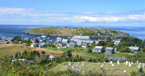 A scenic view of a coastal village with gray buildings, boats in the water, and a grassy landscape under a blue sky.