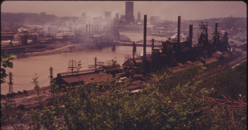 Industrial landscape with smokestacks along a river, city skyline in the background, and greenery in the foreground.