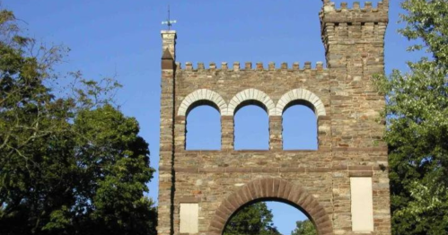 Stone archway with three arched windows, topped with a tower, surrounded by trees and a clear blue sky.