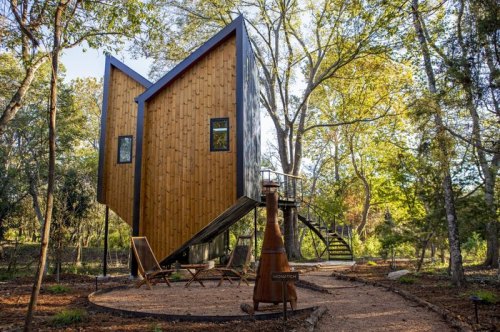 A modern wooden treehouse with a unique design, surrounded by trees and a pathway leading to outdoor seating.