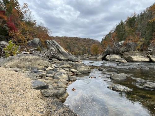 A serene river scene with smooth rocks, sandy banks, and autumn foliage under a cloudy sky.