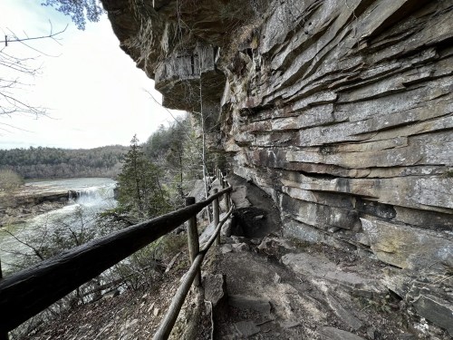 A rocky cliffside path with a wooden railing, overlooking a river and waterfall in a wooded area.