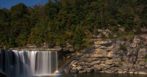 A serene waterfall cascades over rocky cliffs, surrounded by lush greenery and a faint rainbow in the mist.
