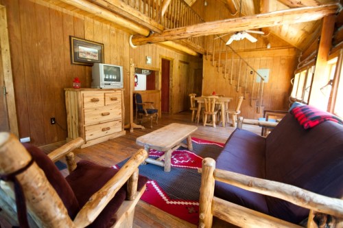 Cozy wooden cabin interior with a sofa, chairs, dining table, and staircase, featuring rustic decor and warm lighting.