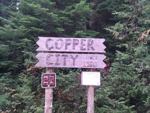 Wooden sign for Copper City, established in 1960, surrounded by dense green trees.