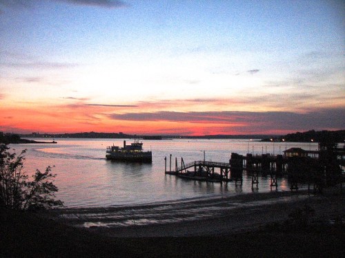A ferry sails on a calm river at sunset, with vibrant colors in the sky and a silhouette of a pier in the foreground.