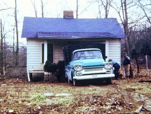 A vintage blue truck parked in front of a small, weathered house in a wooded area, with two people nearby.