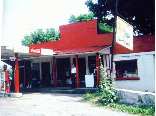 A vintage store with a red exterior, featuring a Coca-Cola sign and a gas pump, surrounded by greenery.