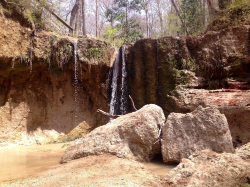 A small waterfall cascades over rocky terrain, surrounded by trees and a calm pool of water below.