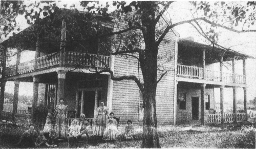 A historic two-story house with a porch, surrounded by trees, with several people sitting in front.