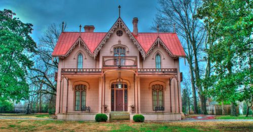 A large, ornate pink Victorian house with a red roof, surrounded by trees and a grassy lawn.