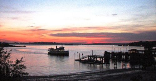 A ferry sails on a calm river at sunset, with vibrant colors reflecting on the water and a dock in the foreground.