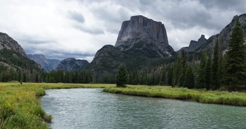 A serene river flows through a lush valley, framed by towering mountains and overcast skies.