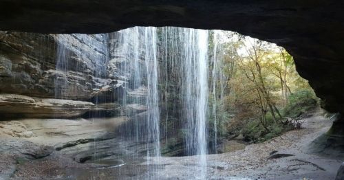 A serene waterfall cascades through a rocky arch, surrounded by lush greenery and autumn foliage.