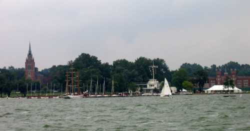 A scenic view of a lake with sailboats, a historic building, and trees under a cloudy sky.