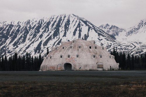 A weathered, dome-shaped structure stands in front of snow-capped mountains and a forested area under a cloudy sky.