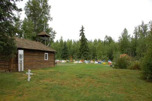 A wooden building beside a grassy area with a white cross, surrounded by trees and colorful tents in the background.