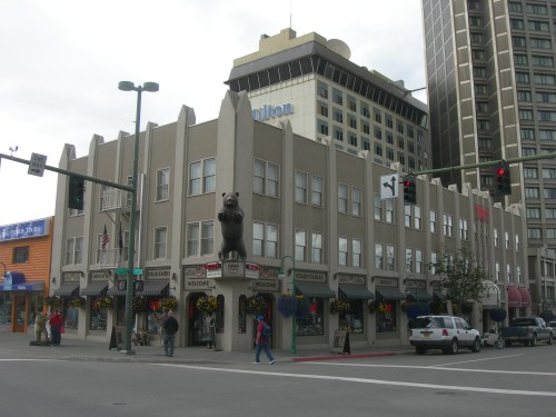 A building with a bear statue on top, surrounded by shops and tall buildings, with traffic lights in the foreground.