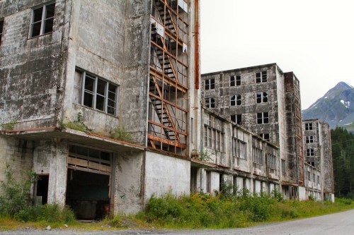 Abandoned industrial buildings with weathered concrete and broken windows, surrounded by greenery and mountains.